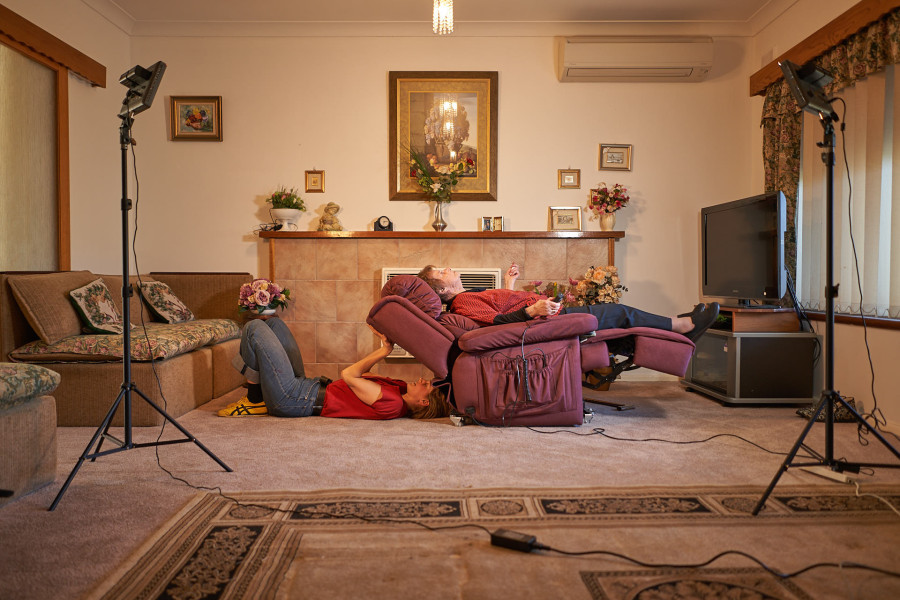 In a lounge room, an older woman reclines on a recliner chair, a younger woman lays on the floor holding up the back of the recliner chair.