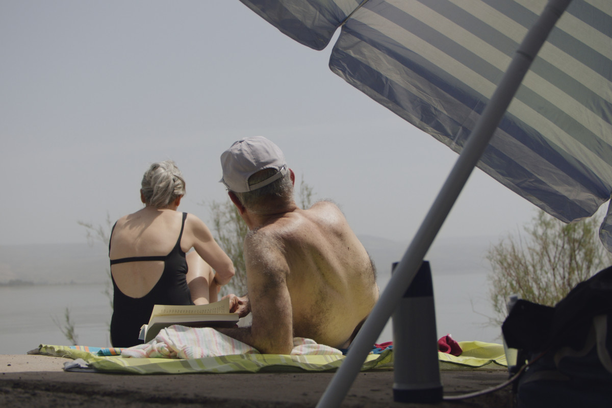 A woman and a man are resting in bathers near a body of water. The man reads a book. There is an umbrella behind them in the front of the frame.