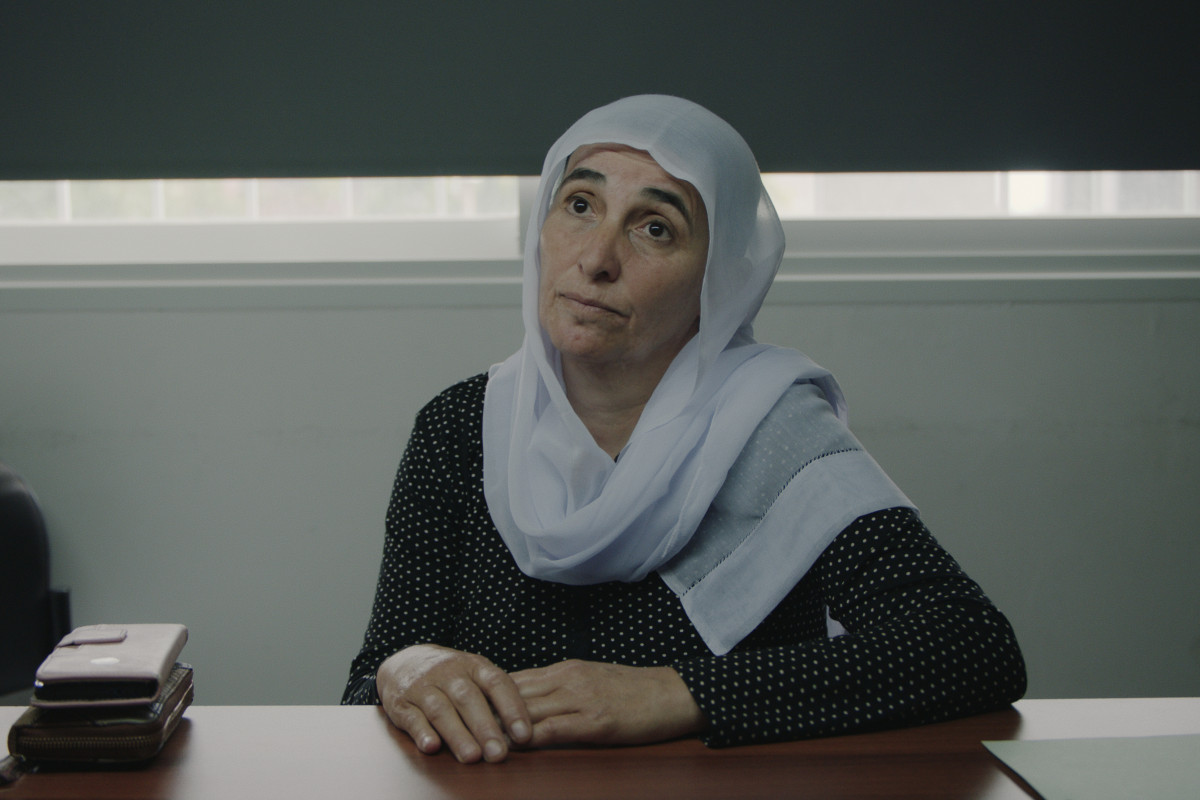 A woman sitting at a table looking upward away from the camera. She has a mostly blank expression. She wears a black spotted long sleeve and a headscarf/Shayla/hijab? A phone and wallet rest beside her