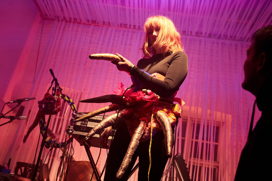 Performer in octopus-like costume holding bread on stage under pink lighting during electronic music performance.