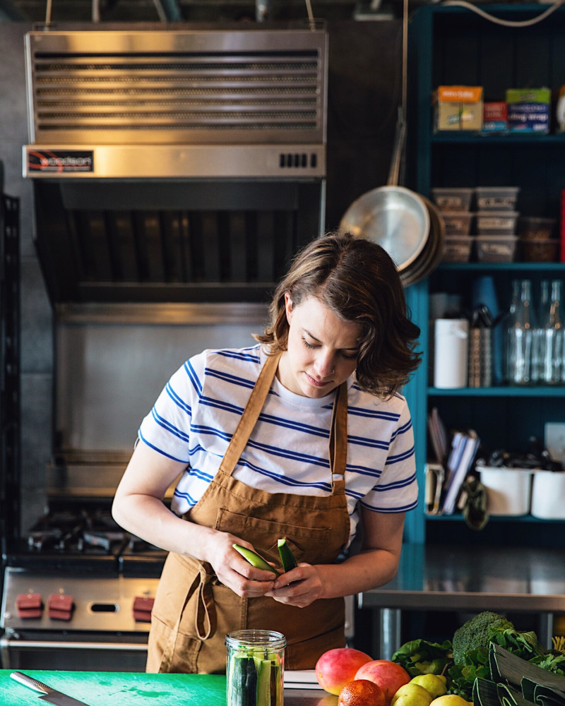 Jessie Spiby stands behind a kitchen counter, working with cucumbers and various vegetables.