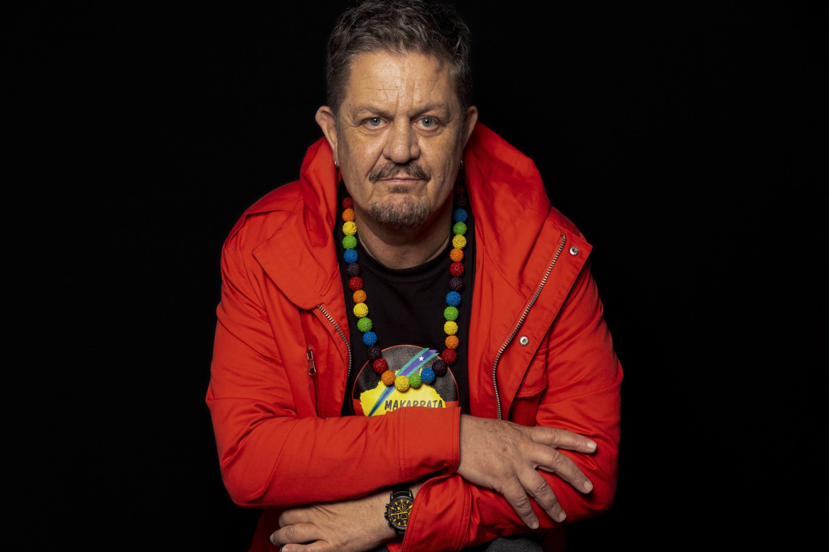 Peter Waples-Crowe has arms crossed, looks to camera. He wears a red jacket, and rainbow beaded necklace.