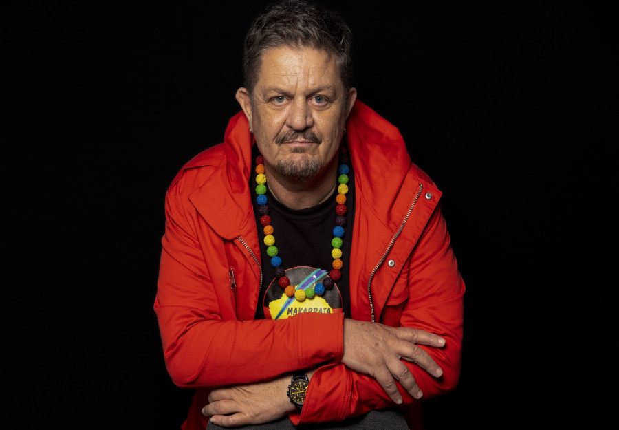 Peter Waples-Crowe has arms crossed, looks to camera. He wears a red jacket, and rainbow beaded necklace.