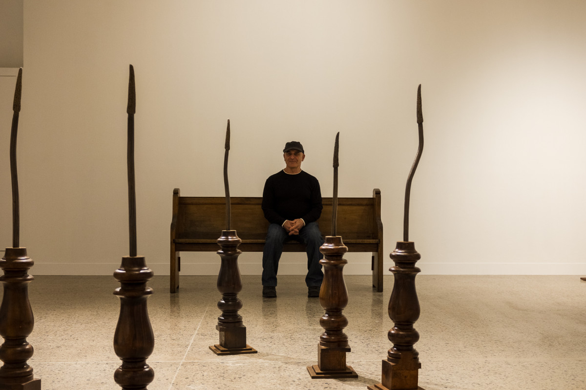 Lee sits on a church pew smiling to camera. 5 artworks - brass cast xanthorrhoea's on wooden table legs are visible.