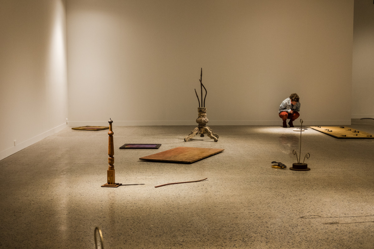 Installation view of Lee's work in the gallery. Artworks are scattered across the floor. One person is kneeling down looking at a particular artwork on the gallery floor.