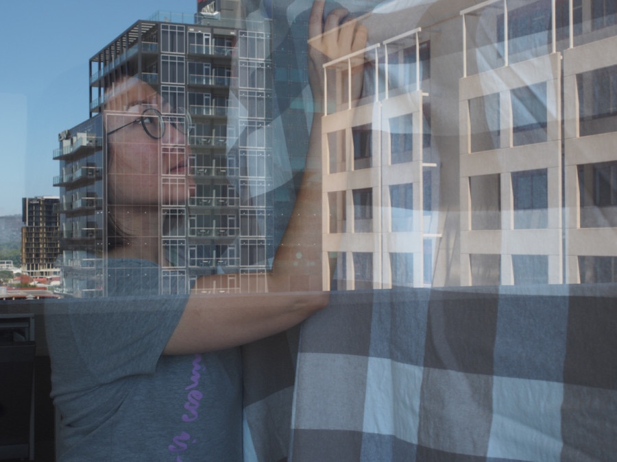 A double exposure image showing a person indoors adjusting a large checkered fabric, overlaid with the reflections of modern apartment buildings in a glass window. Luna Chan's face, partially obscured by the reflections, appears thoughtful and focused. The composition blends interior and exterior spaces, creating a layered urban scene.