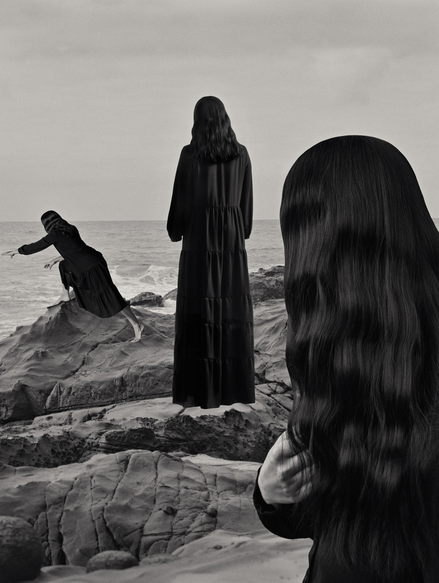 Black and white artwork of three cloaked figures on rocks on the beach.