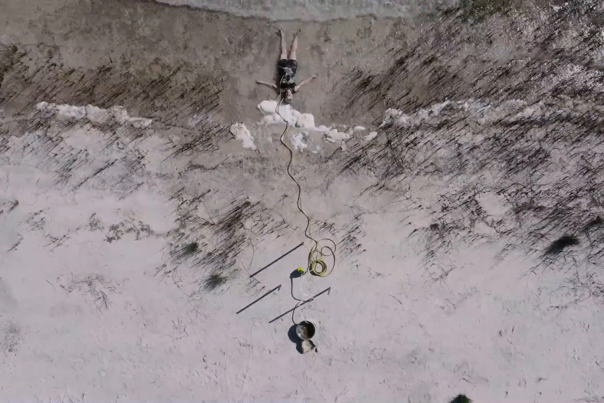 A person lies on a sandy beach near the shoreline, connected to a long yellow cord leading to objects in the sand.