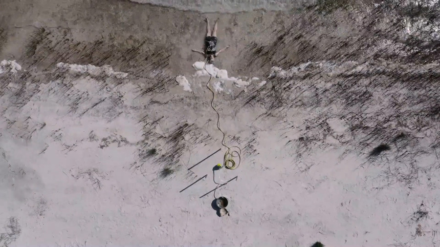 A person lies on a sandy beach near the shoreline, connected to a long yellow cord leading to objects in the sand.