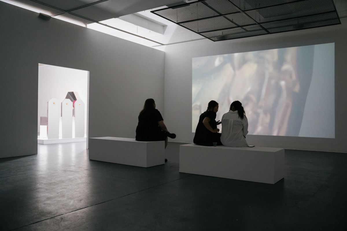 Opening night. 2 people sit on one plinth and 1 person sits on the other plinth to watch Georgia Button's moving image artwork projection. Wide angle view.