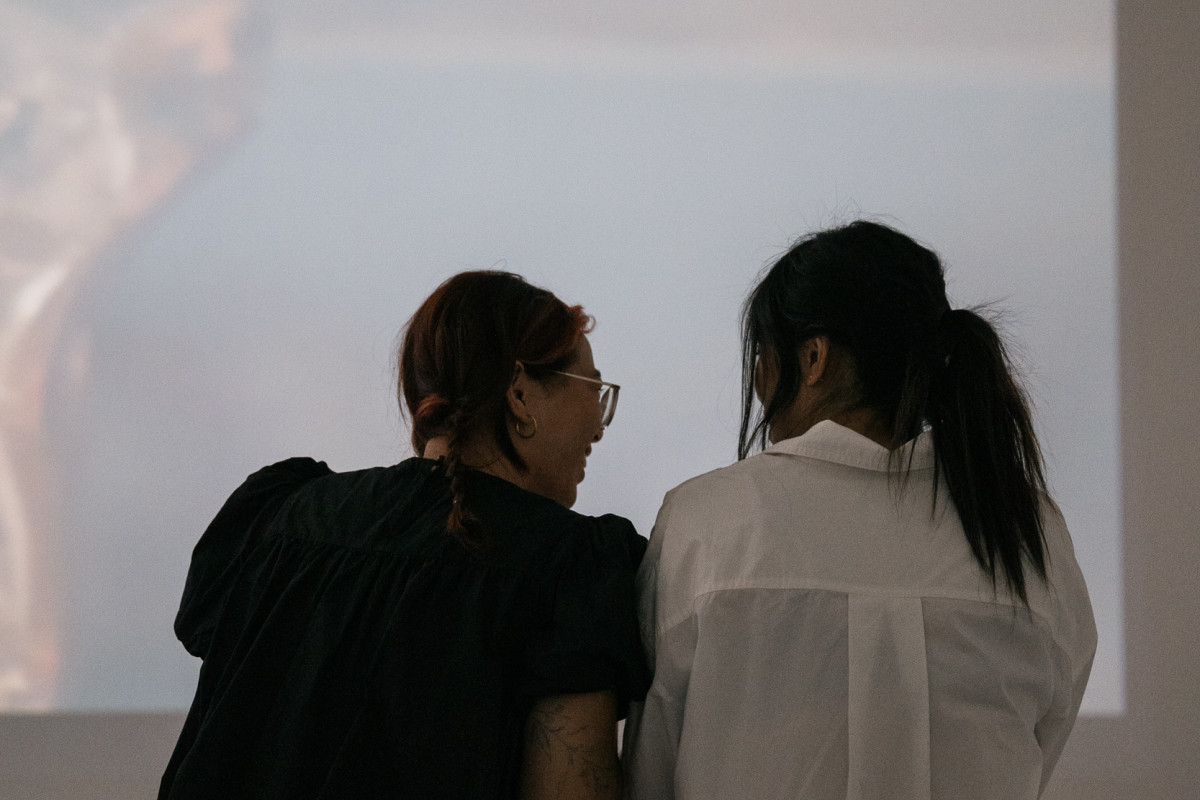 Opening night. 2 people lean together smiling and watching Georgia Button's moving image artwork projection.