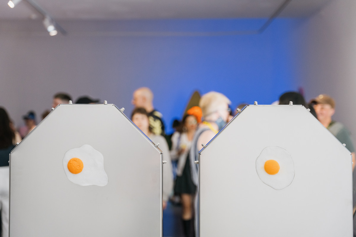 Close up of Jennifer's work - the back of 2 metal chairs with resin cast fried eggs in the centre of each. The opening night audience gather around and are blurred in the background.