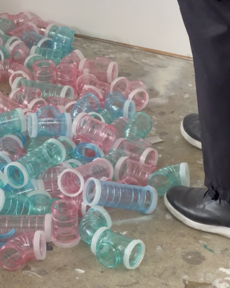 A pile of pink, blue and green transparent tubes lay on Emmaline Zanelli's art studio floor. A person stands to the right, wearing dark navy pants and black shoes.