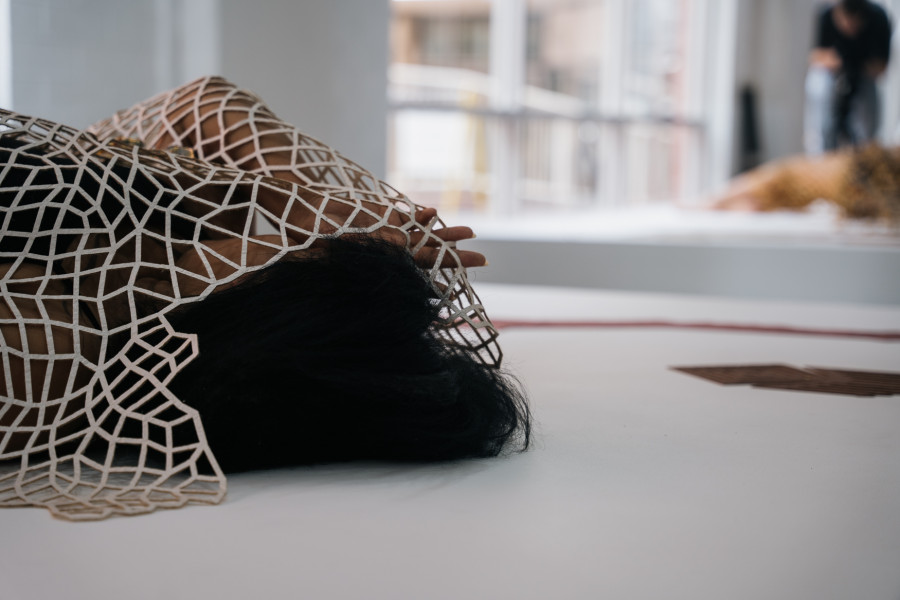A person lying on a white surface with a geometric net-like structure covering their body and hair. In the background, another figure is visible in a bright gallery space with large windows.