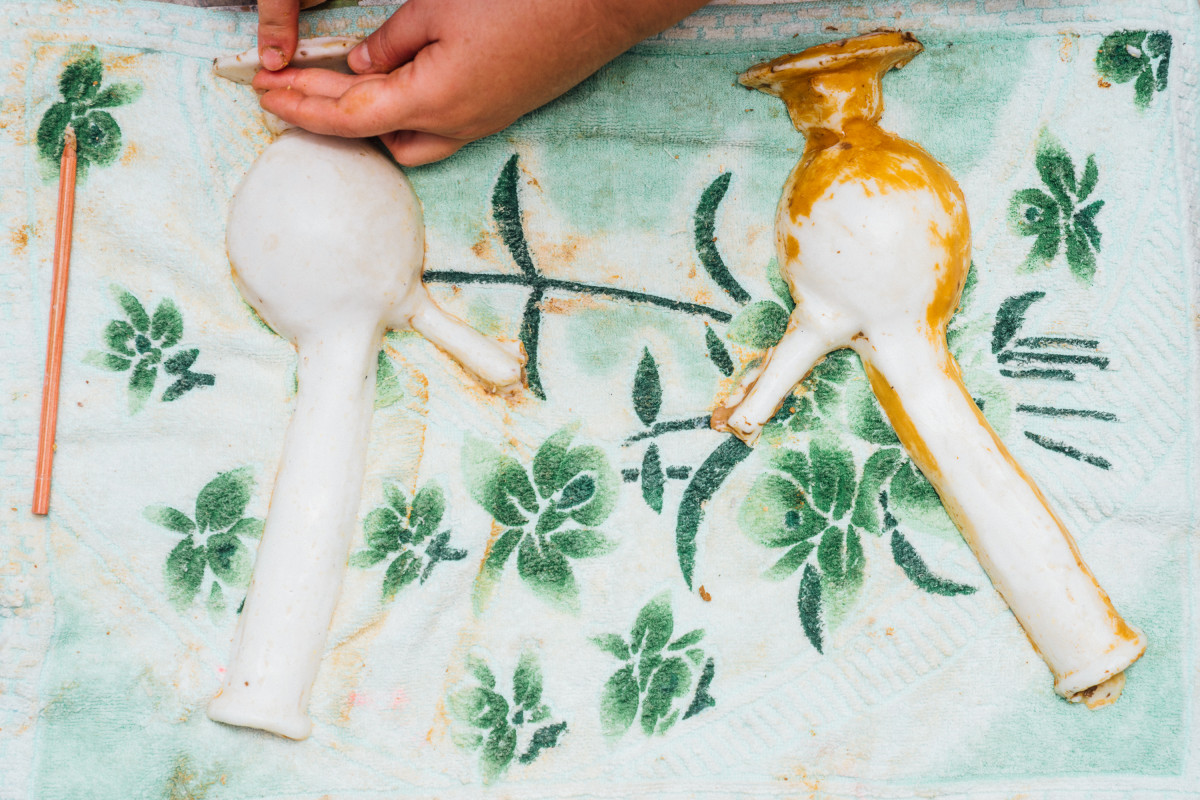 Two white soap casts of a bong rest on a green towel with a floral pattern. The artist Teresa Busuttil’s hands touch the base of the soap bong. A pencil rests on the towel on the left.