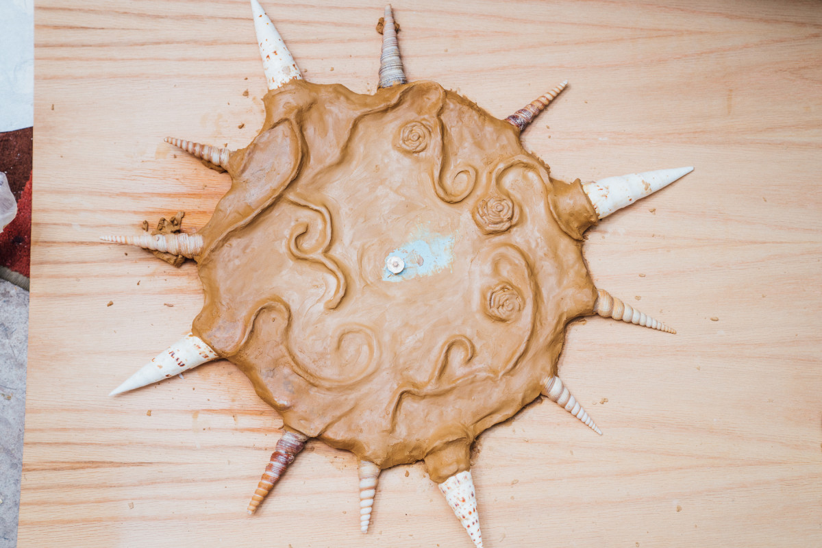 A clock work-in-progress artwork rests on plywood. The clock is made of clay, is framed by 12 pointy shells and features swirl and rose patterns on the face.