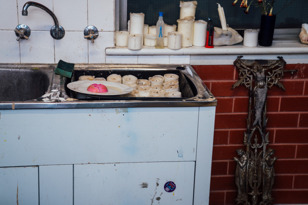 Inside Teresa Busuttil’s Studio there is a sink and a window sill. Used candle pillars fill the right sink trough and the window sill. A large ornate religious crucifix rests against the brick wall under the window sill.