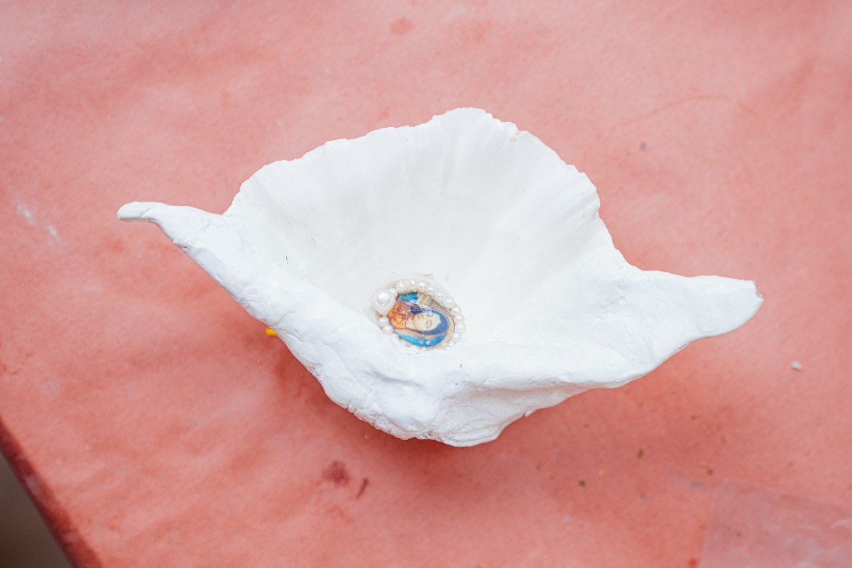 A white shell with a fanned shape sits on a corner of a table which is covered in butcher's paper. The shell’s inside faces up. A small oval portrait of Virgin Mary framed by small pearls is adhered in the centre of the shell.