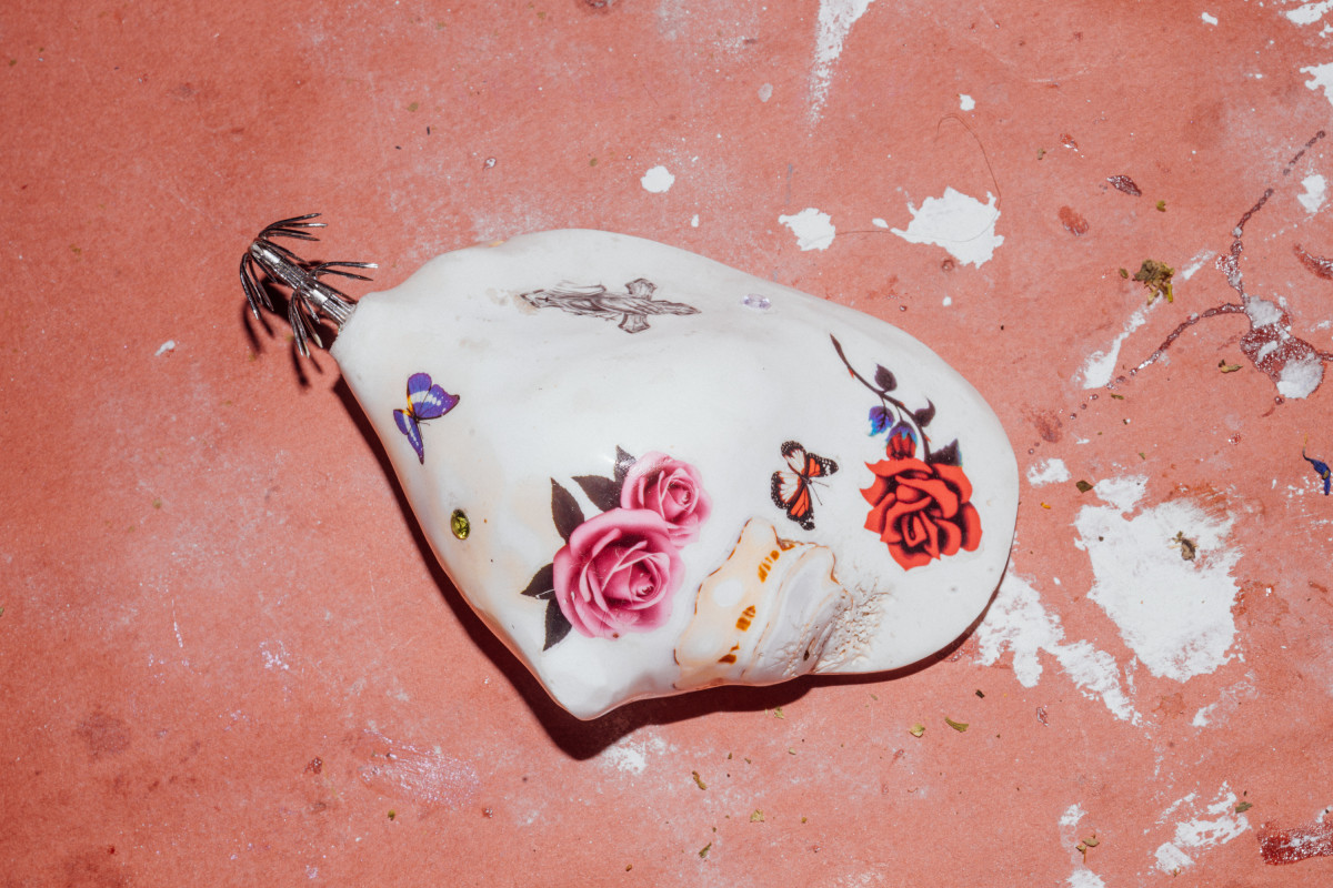 A large white and smooth shell sits on a surface with paint splatters on it. Several coloured temporary tattoos and two rhinestones are on the outside of the shell. A fishing hook is adhered to the end of the shell.