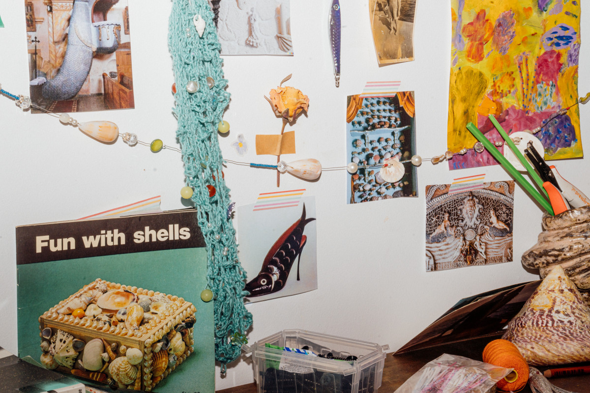 The wall of Teresa Busuttil’s Studio. Several colourful posters hang on the wall, as well as a netted crochet bag, a dried rose, a purple fishing lure, and bunting made of shells and beads. A book titled ‘Fun with shells’ rests against the wall on a table. A large conical shell, a thread spool and a filled pencil pot sit on the table.