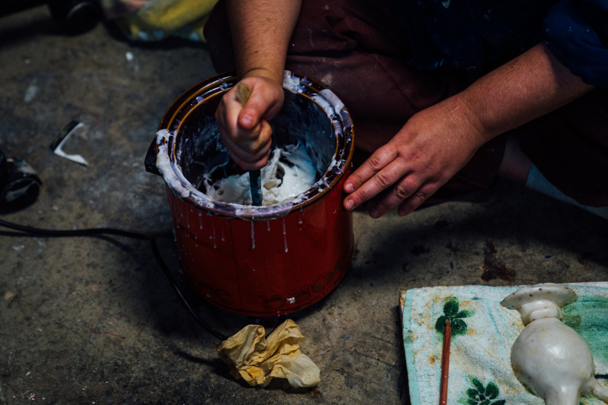 Teresa Busuttil works on the floor of her studio. You can only see her hands which are stirring melted wax in a small and red candle melting pot. A scrunched piece of paper towel sits in front of the pot. In the bottom right corner you can see the bottom of her work-in-progress, a candle cast bong.