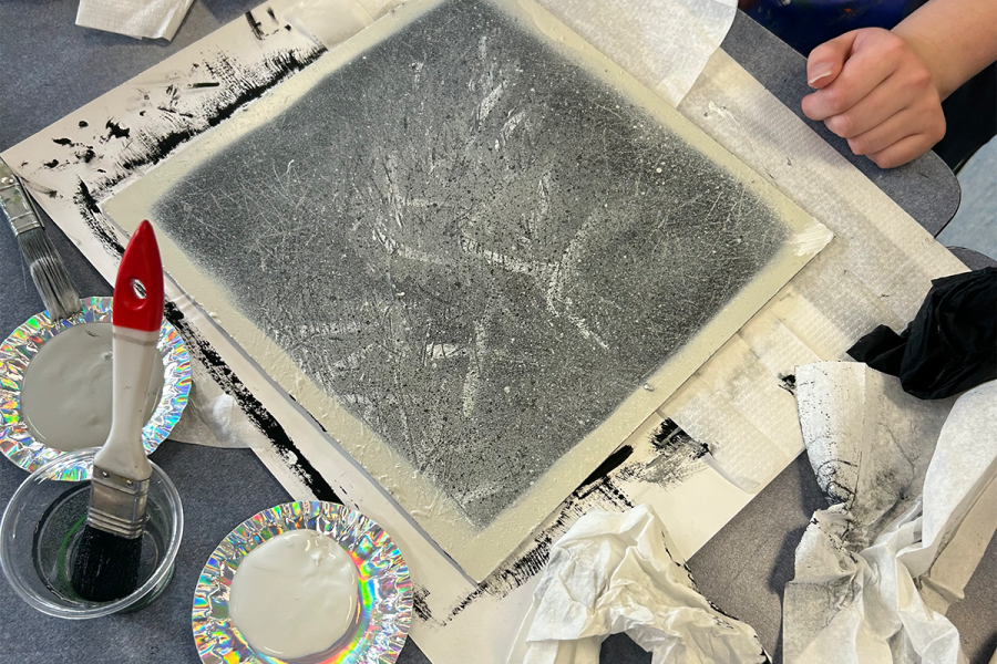 Student artist experimentation from Thomas Readett workshop. A square piece of cardboard with with grey paint splatters. 2 small silver plastic plates hold paint - grey and white. A paintbrush sits in a plastic cup of water.