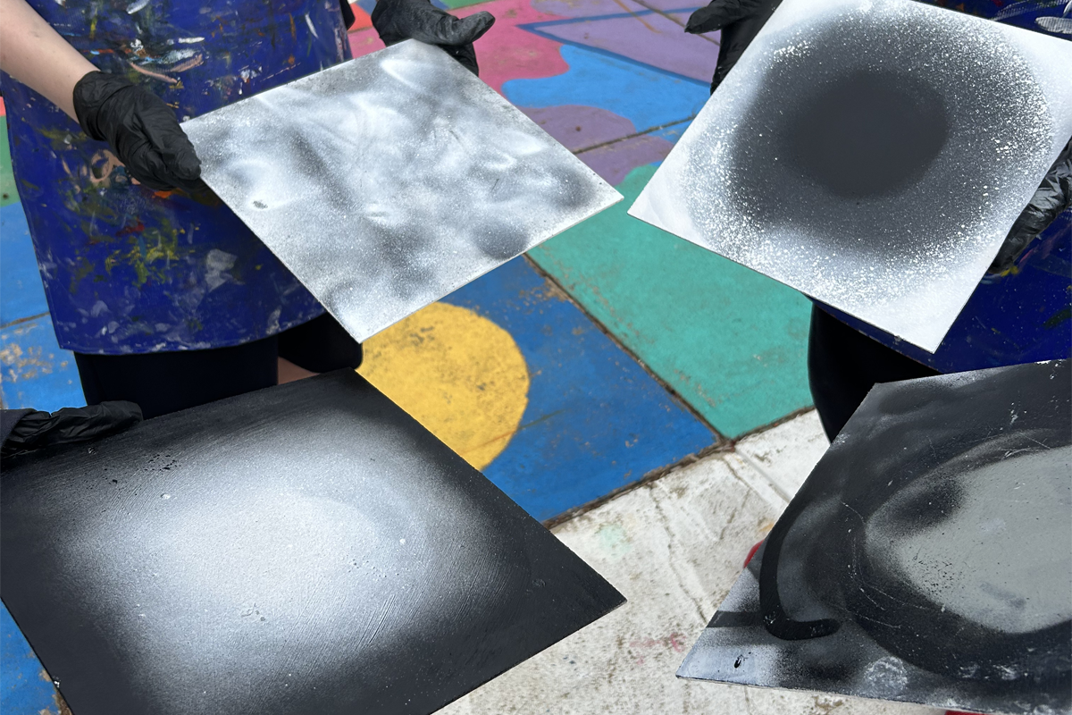Student artist experimentation from Thomas Readett workshop. 4 students hold their artworks in a circle. 4 pieces of cardboard with various grey white and black spray painted works.