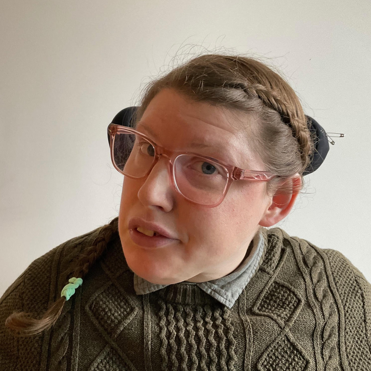 I'm 39 years old. I have mousey brown braided hair in the style of Katniss Everdeen and fair skin. I’m wearing plain clear pink framed glasses, a grey shirt and a green jumper. I'm looking at the camera like I know everything with a slight smile.