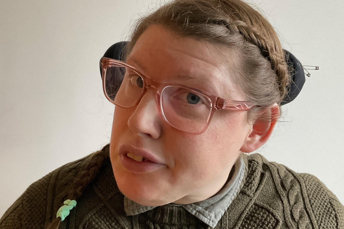 I'm 39 years old. I have mousey brown braided hair in the style of Katniss Everdeen and fair skin. I’m wearing plain clear pink framed glasses, a grey shirt and a green jumper. I'm looking at the camera like I know everything with a slight smile.