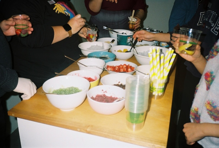 A group of people gathered around a wooden table filled with bowls of colourful ingredients, assembling halo-halo drinks.