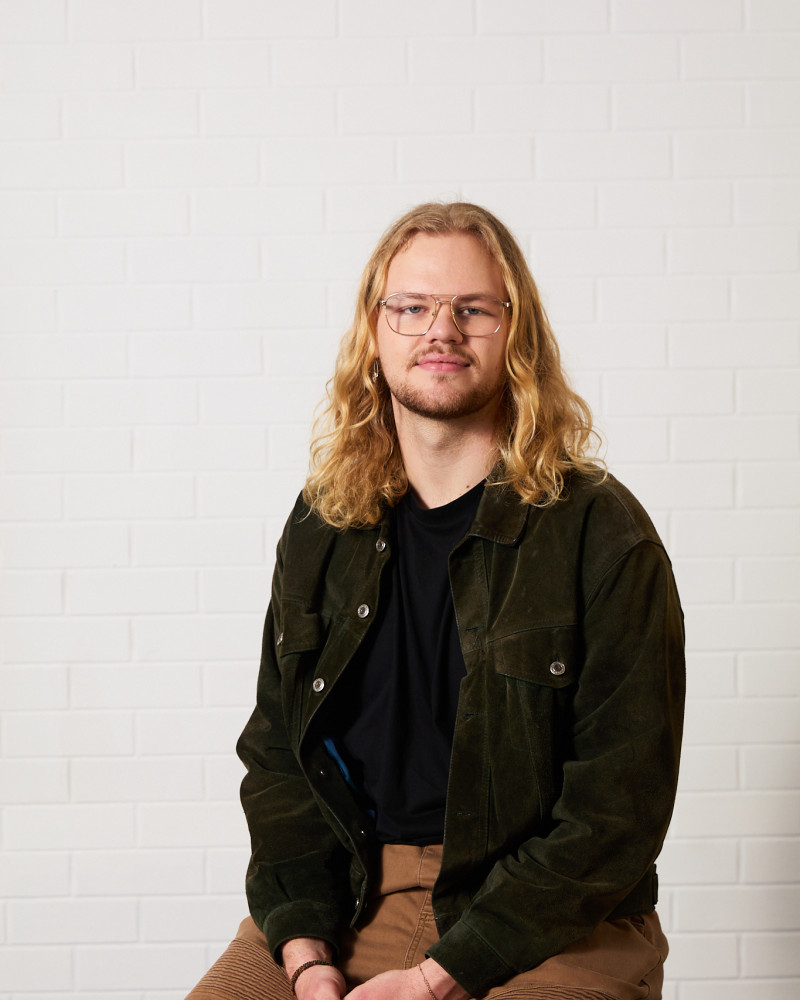 Headshot of William Maggs. William has wavy blonde shoulder length hair, blue eyes and blonde facial hair. They are wearing a dark green suede jacket, black t-shirt, and blown pants. They sit against a white background, smiling directly at the camera.