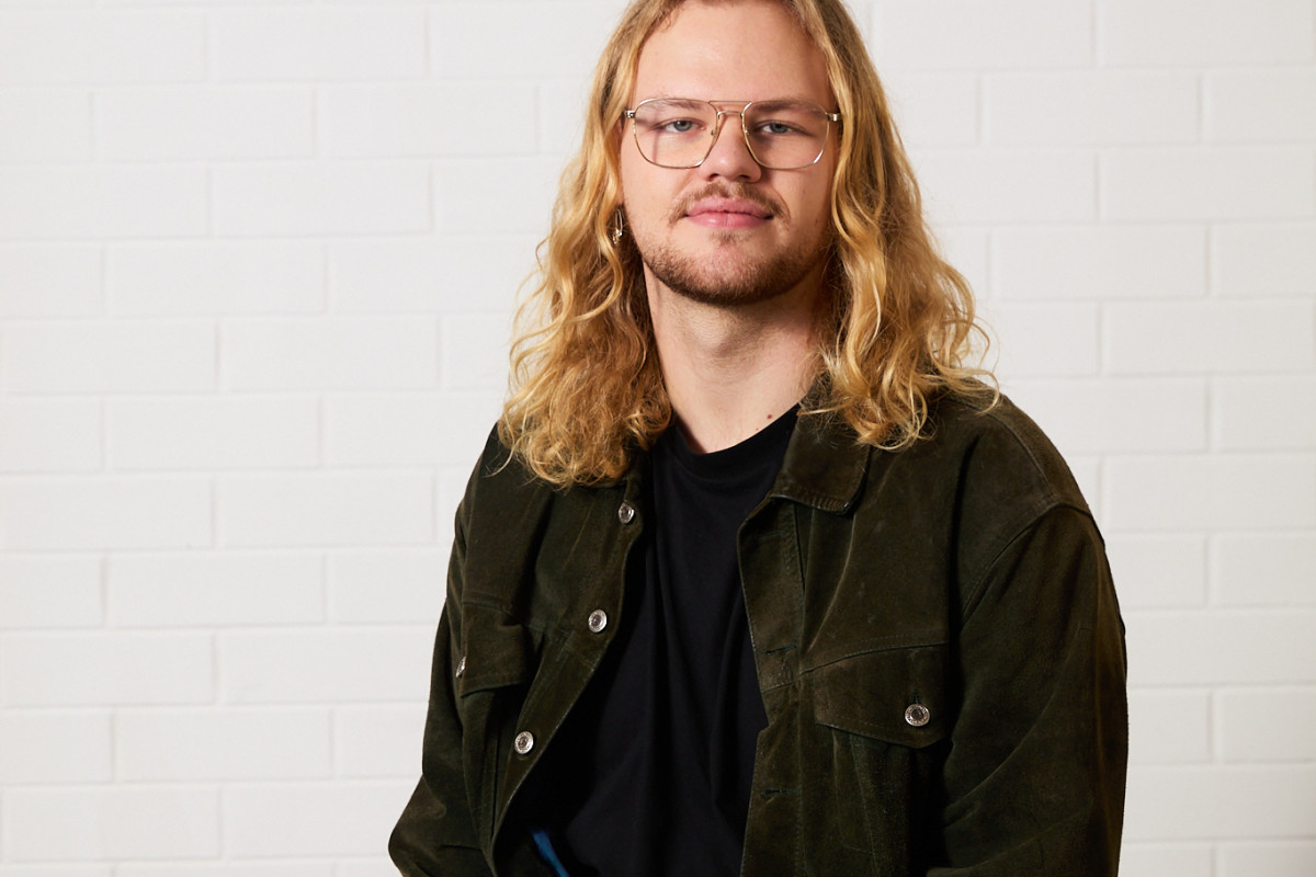 Headshot of William Maggs. William has wavy blonde shoulder length hair, blue eyes and blonde facial hair. They are wearing a dark green suede jacket, black t-shirt, and brown pants. They sit against a white background, smiling directly at the camera.