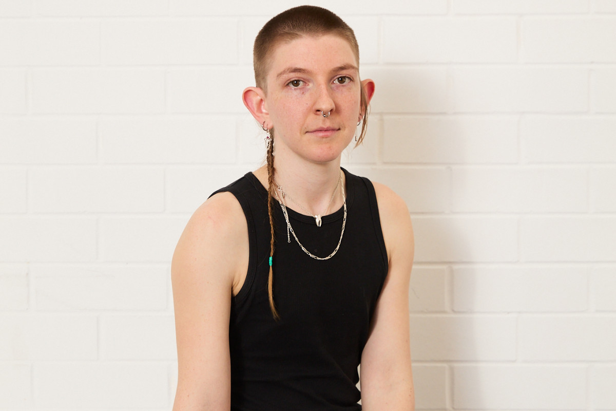 Headshot of Grace Marlow. Grace has pale white skin, brown eyes, and a silver septum nose ring. They are wearing a black tank top and black pants. They have a blonde buzz cut and a thinly braided 'rat's tail', which is draped over their right shoulder. They sit against a white background, looking directly at the camera.