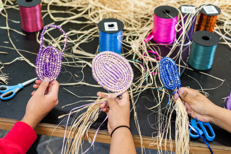 Three hands hold brightly coloured weavings over a black workshop table that has various materials laid out - scissors, thread, ribbon.