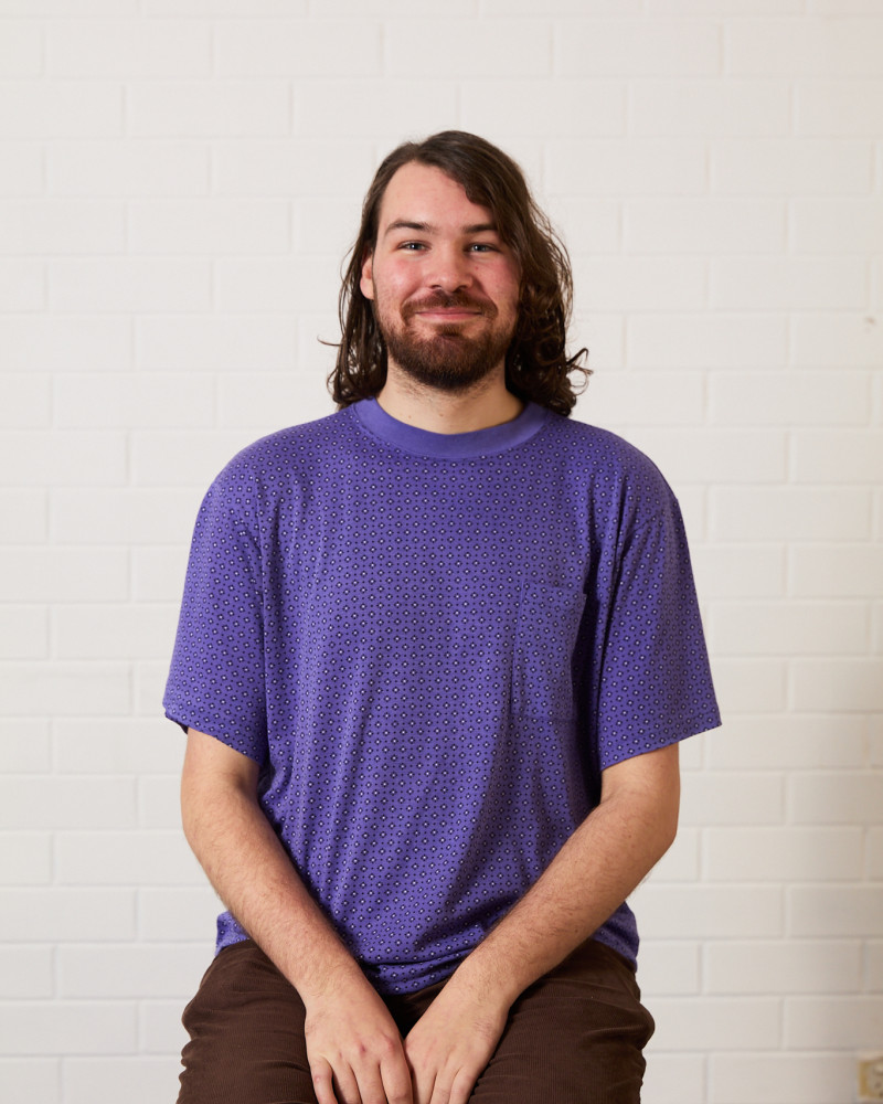 Headshot of Hen Vaughan. Hen has dark shoulder length hair, blue eyes and brown facial hair. They are wearing a purple patterned t-shirt and chocolate brown jeans. They sit against a white background, smiling directly at the camera.