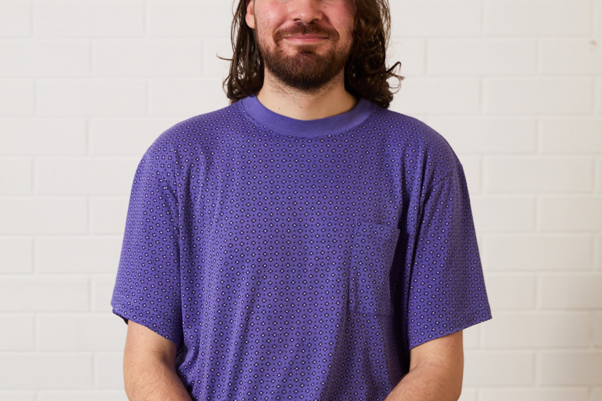 Headshot of Hen Vaughan. Hen has dark shoulder length hair, blue eyes and brown facial hair. They are wearing a purple patterned t-shirt and chocolate brown jeans. They sit against a white background, smiling directly at the camera.