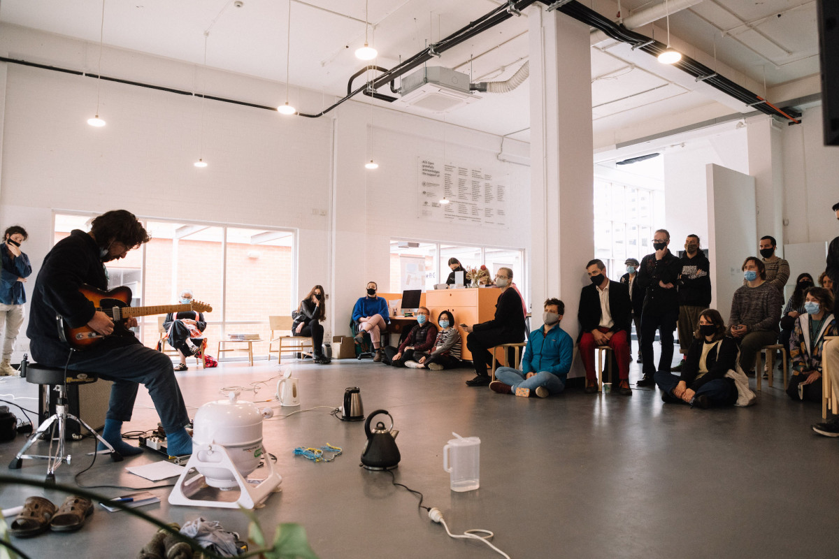 Musician plays guitar for audience. Kitchen appliances are displayed on the floor before him.
