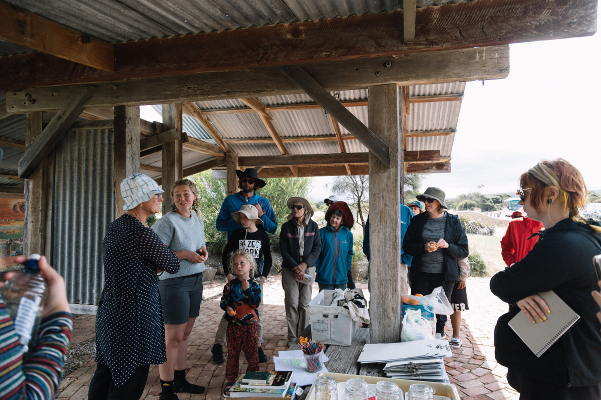 Group gathers under shelter. Clip-boards and pencils and books are on the tabel.
