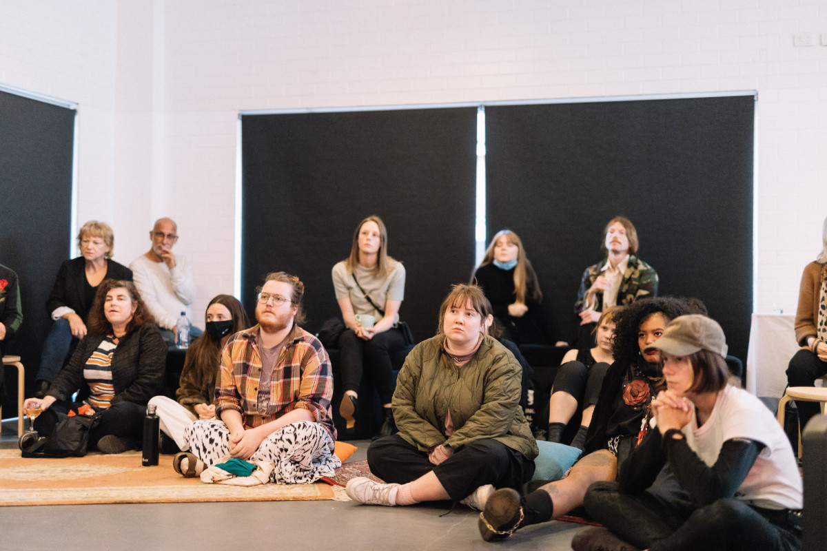 Audience sits on ground in front room, watching intently.