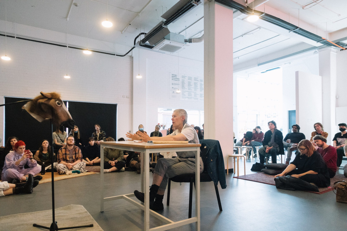 A person sits at a table facing a horse head mask. Audience watches.