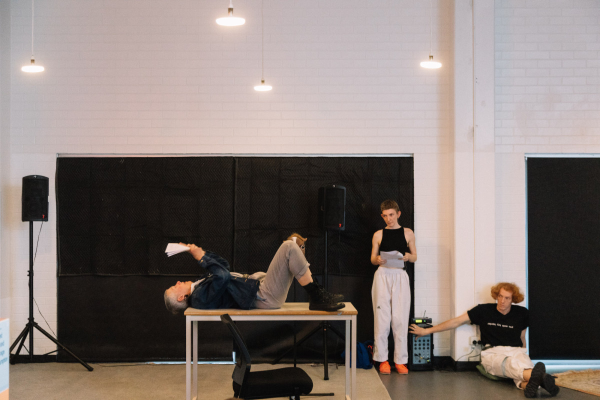A person lies on their back on a table, reading from paper. Audience watches.