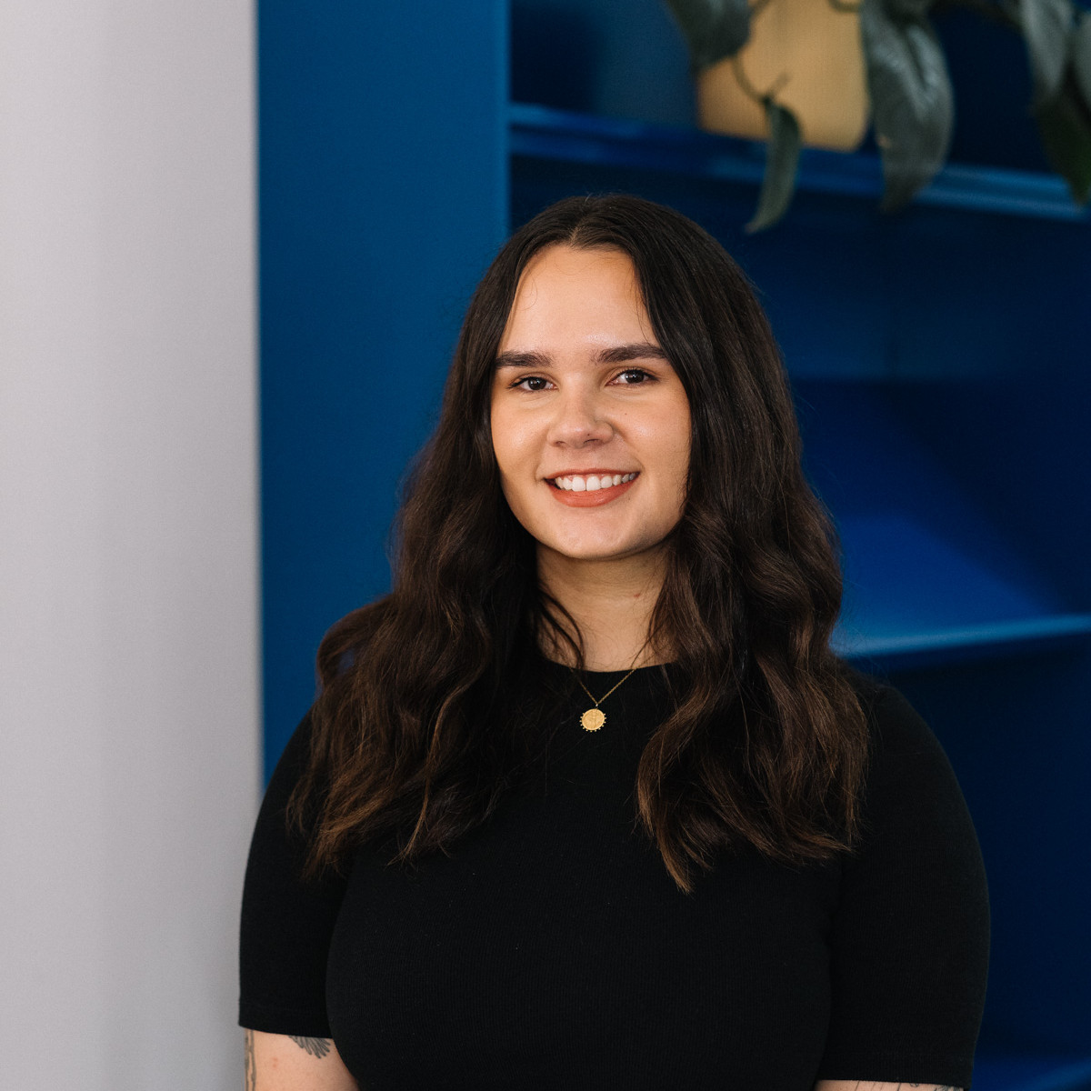 Carly Tarkari Dodd headshot. Dodd smiles to camera wearing all black. She is standing in front of a blue bookcase.