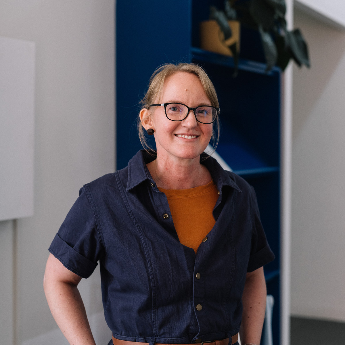 Marian Sandberg headshot. Sandberg smiles to camera, she is standing in front of a blue bookcase. Marian wears blue overalls and black glasses.