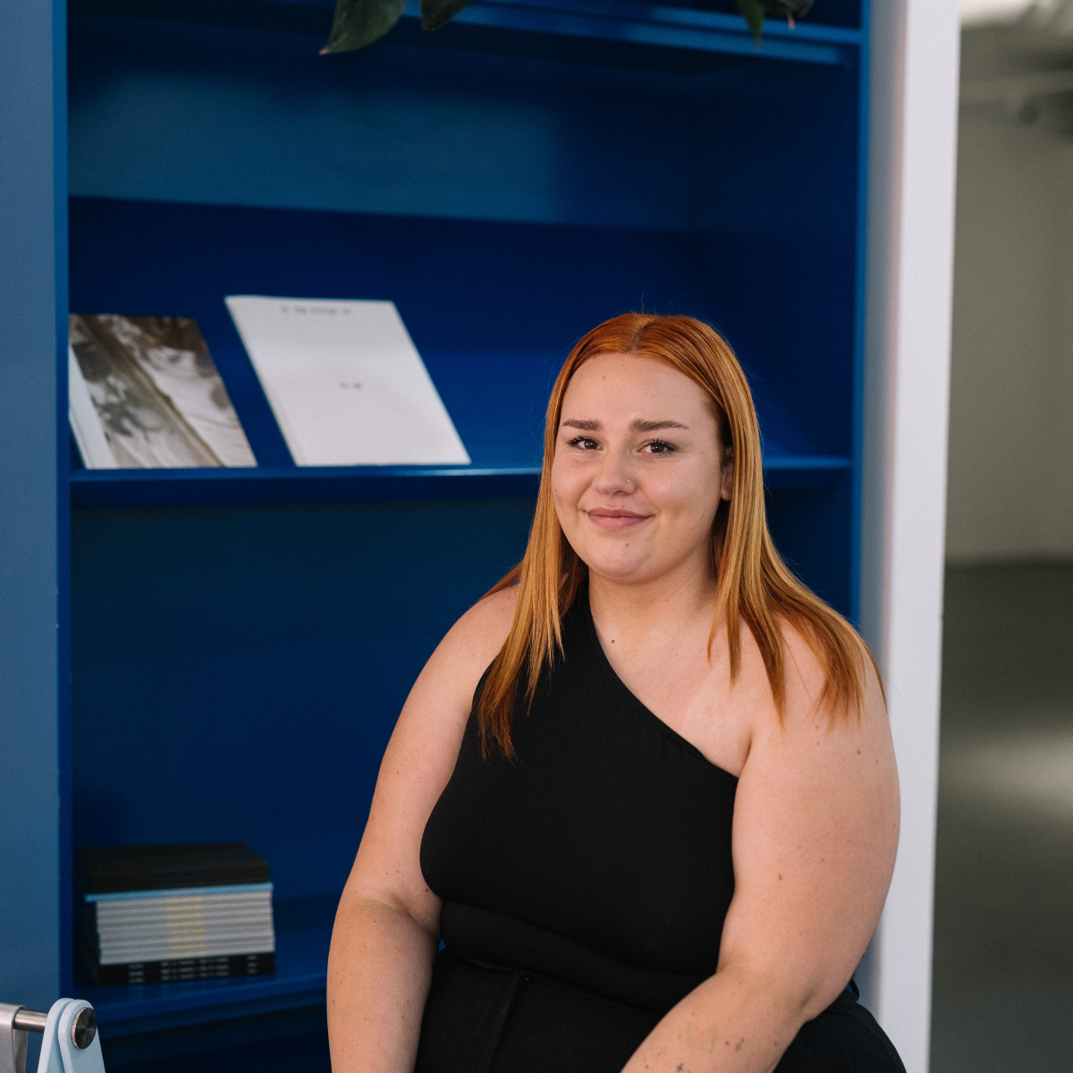 Abbey Murdoch headshot. Murdoch smiles to camera wearing all black. She is sitting in front of a blue bookcase.