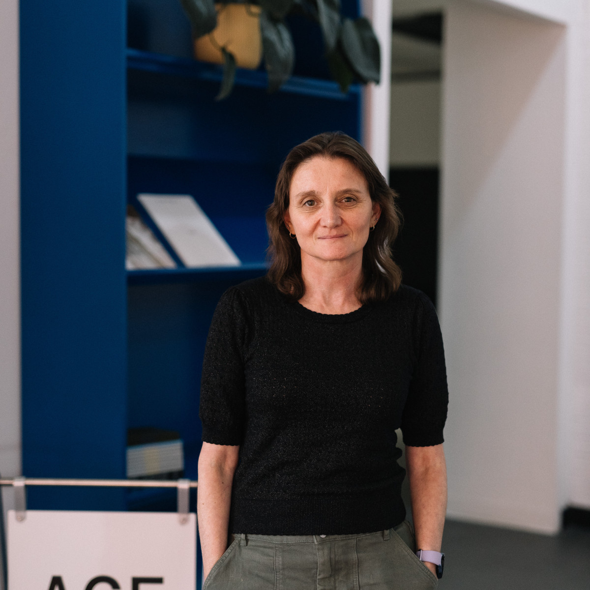 Katey Smoker headshot. Smoker looks to camera wearing a black top and a khaki skirt. She is standing in front of a blue bookcase.