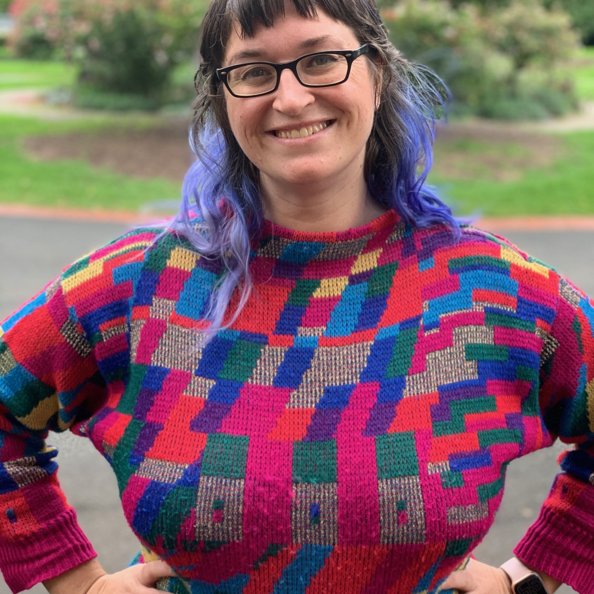 Headshot of Elizabeth Reed. I have dyed brightly coloured hair and thick black glasses. I am wearing a geometric-patterned rainbow jumper and I am standing outside in a sunny park.