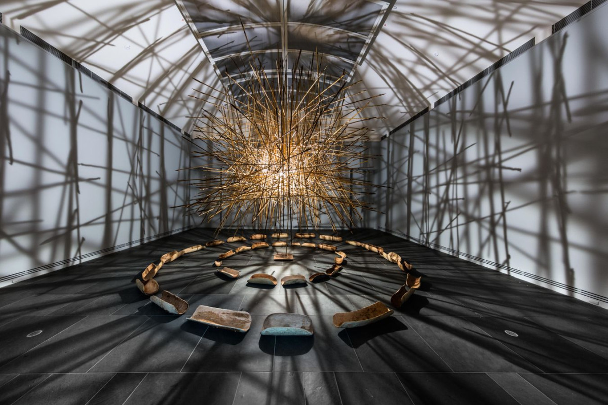 Large installation in centre of room. Light illuminates from within the large hanging spherical stick object. Shadows are cast on walls, ceiling and floor.